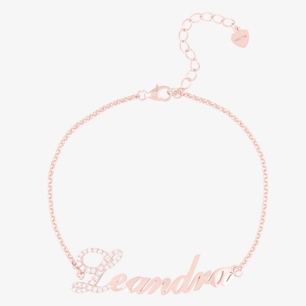 Personalized name bracelet in rose gold with crystal embellishment on first letter, showcasing the name "Leandra" with a delicate chain and heart charm.