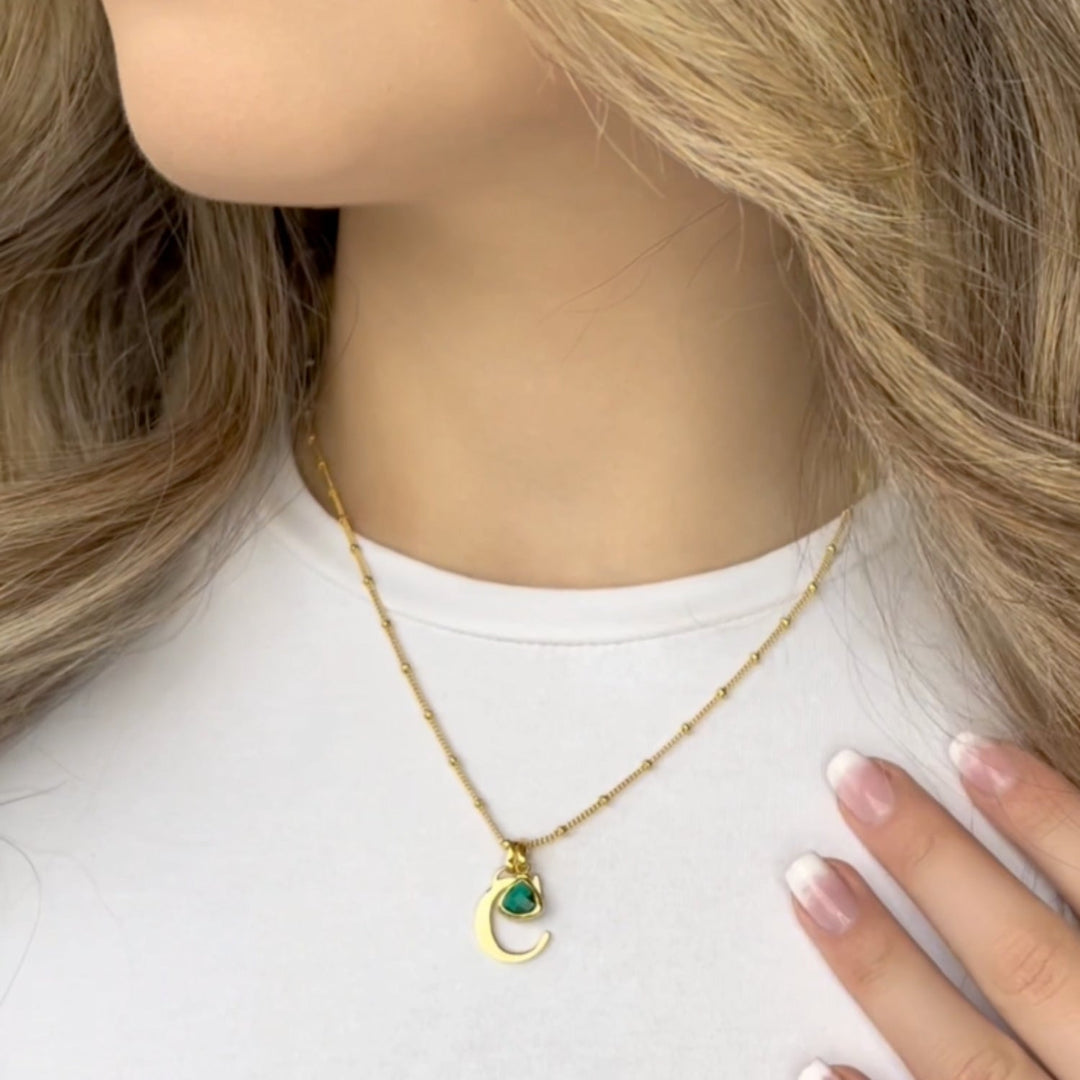 Woman wearing a gold initial necklace with a green birthstone pendant, showcasing elegant jewelry customization options.