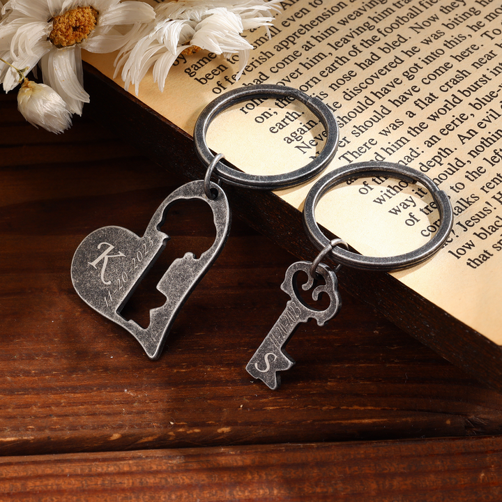 Personalized couple's keychain set with dark grey heart and key charms, featuring custom engraving on wooden background and flowers.