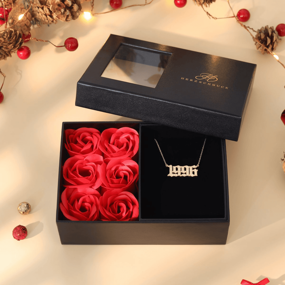 Elegant rosebox with red roses and a gold necklace featuring "1996", presented in a classy black gift box with pinecone and red berry accents.