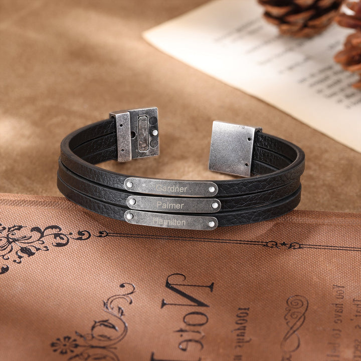 Customizable dark grey leather bracelet with three personalized engravings displayed on a rustic background.