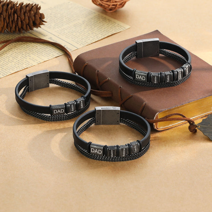 Personalized black leather bracelets with stainless steel chain and 'DAD' engravings on a brown notebook.