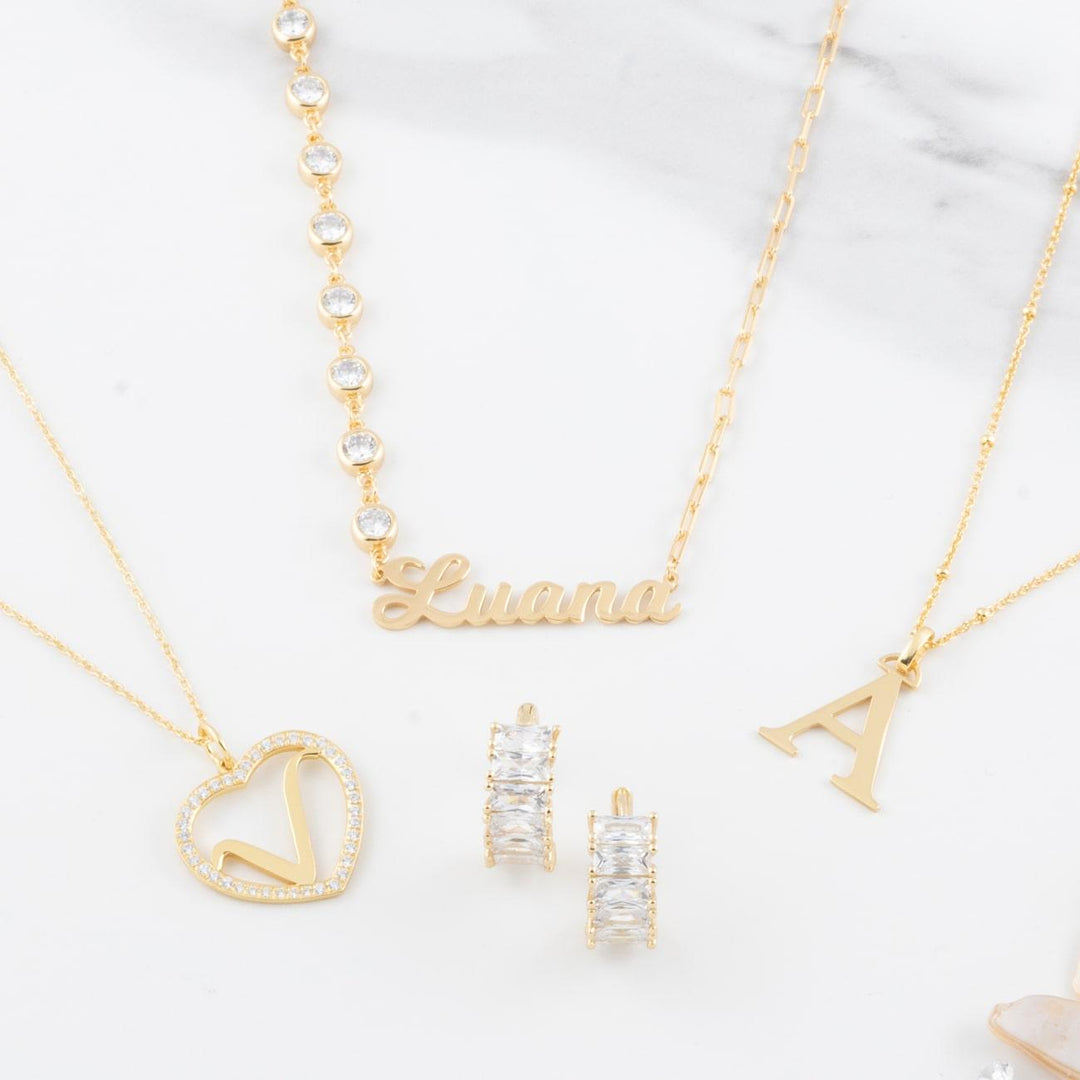 Gold cursive name necklace with zirconia detail, heart pendant, initial pendant, and zirconia earrings on a marble background