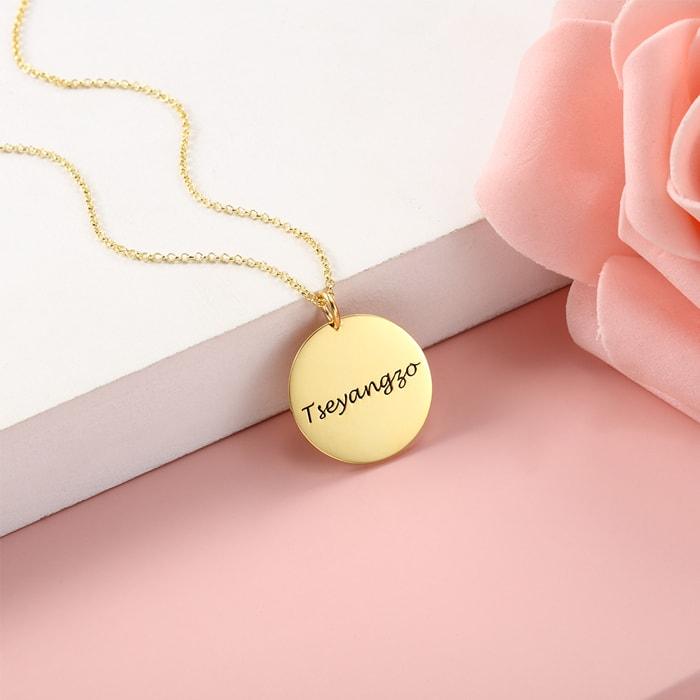 Personalized round medallion pendant necklace in gold with engraving option, displayed on a delicate gold chain, next to a pink rose.