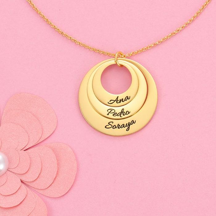 Triple ring engraved pendant necklace in gold with personalized names, displayed on a pink background next to paper flower decoration