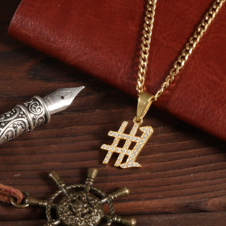 Personalized crystal-encrusted number necklace in gold on a men's curb chain, displayed next to a vintage pen and leather notebook.