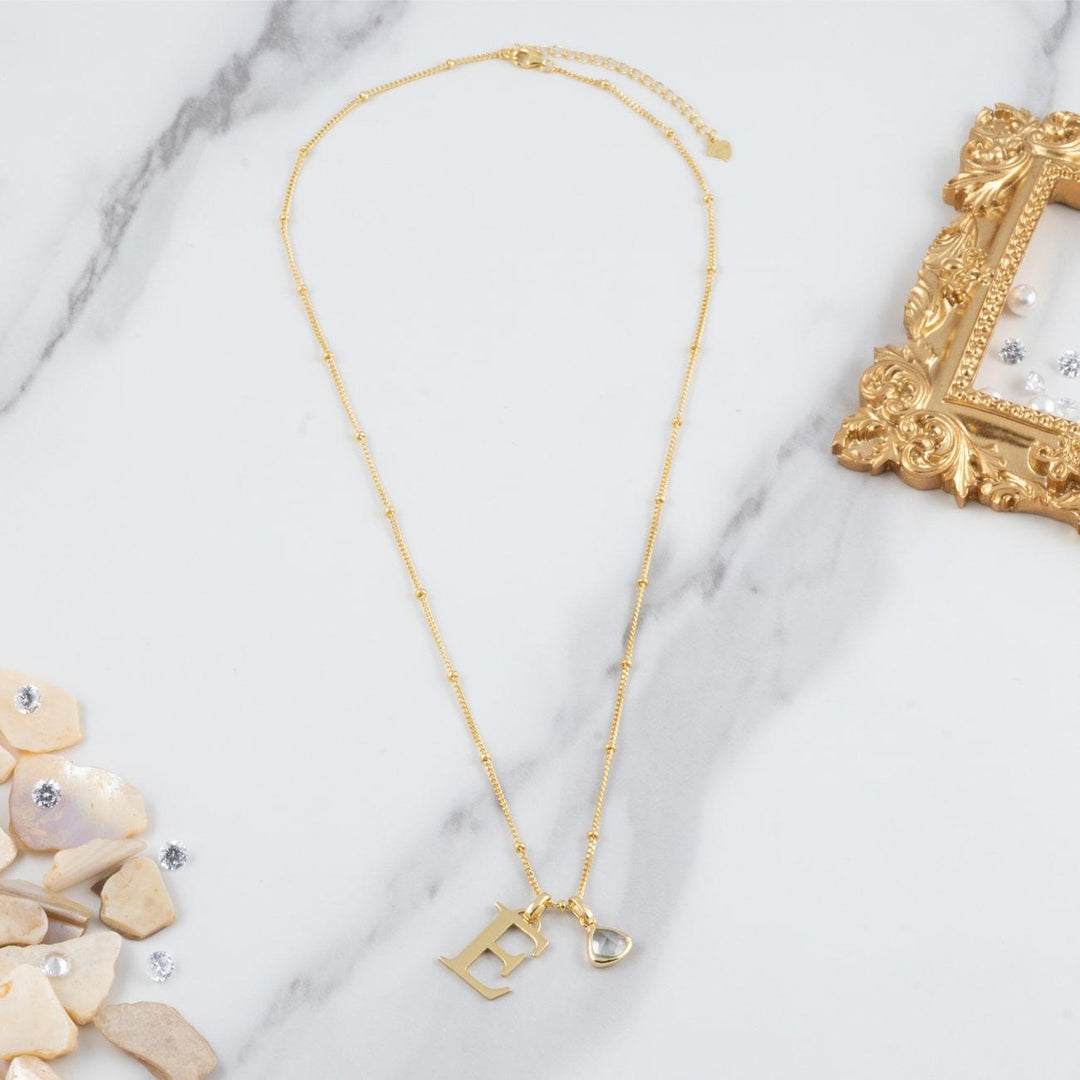 Gold personalized letter necklace with birthstone pendant on white marble background, featuring elegant design and 18K double-plated material.